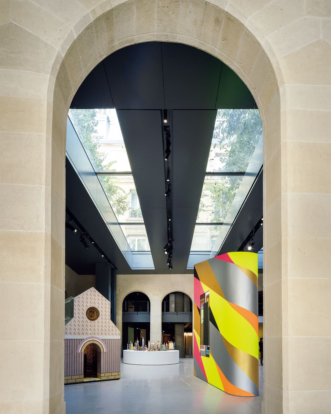 エントランスを入った最初の空間の「建築という装置」と題されたエリア。天窓の上には空と木々が広がり、自然光が展示室にふりそそぐ。右の蛍光色の作品と左のチャペルは、2014年に〈カルティエ現代美術財団〉で展示されたイタリアのデザイナー、アレッサンドロ・メンディーニの作品《OMG!》（2014年）と《Petite Cathédrale》（2002年）。